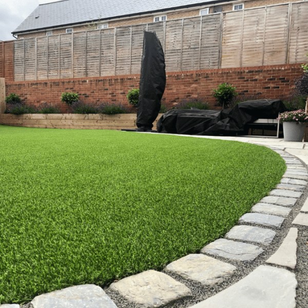 A freshly installed artificial grass lawn edged with curved paving stones in a contemporary garden, with a wooden fence and brick wall backdrop. Block Paving